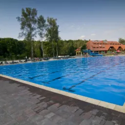 Rozległy, błękitny basen rekreacyjny z wyraźnymi liniami torów, otoczony leżakami i zielenią, z budynkiem w tle. Ujęcie z poziomu chodnika z kostki.