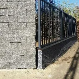 Fragment ogrodzenia z szarej kostki betonowej i metalowych prętów, widok z bliska na połączenie elementów, ziemiste podłoże