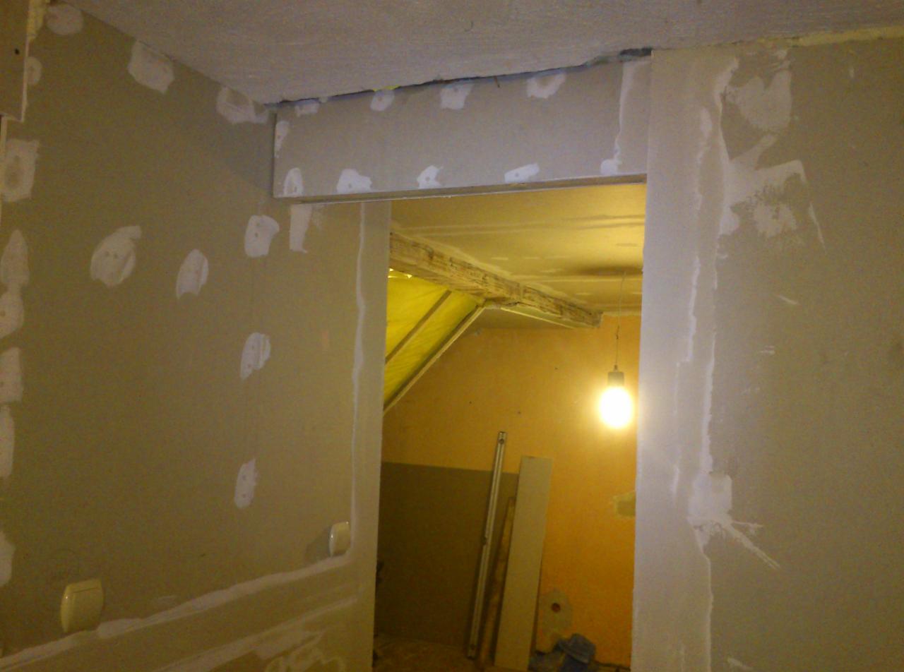 Wnętrze w trakcie prac wykończeniowych: ściany częściowo pokryte gładzią, widoczne szpachlowane miejsca i surowe wykończenie otworu drzwiowego. W głębi pomieszczenia widoczna żarówka.