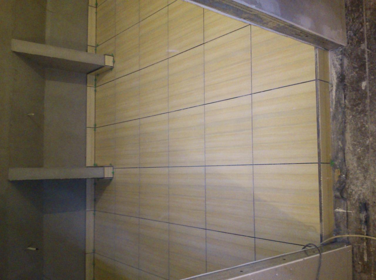 Ułożone beżowe płytki na ścianie z półkami w łazience, widoczne krzyżyki dystansowe i poziomica, podkreślające precyzję wykonania.