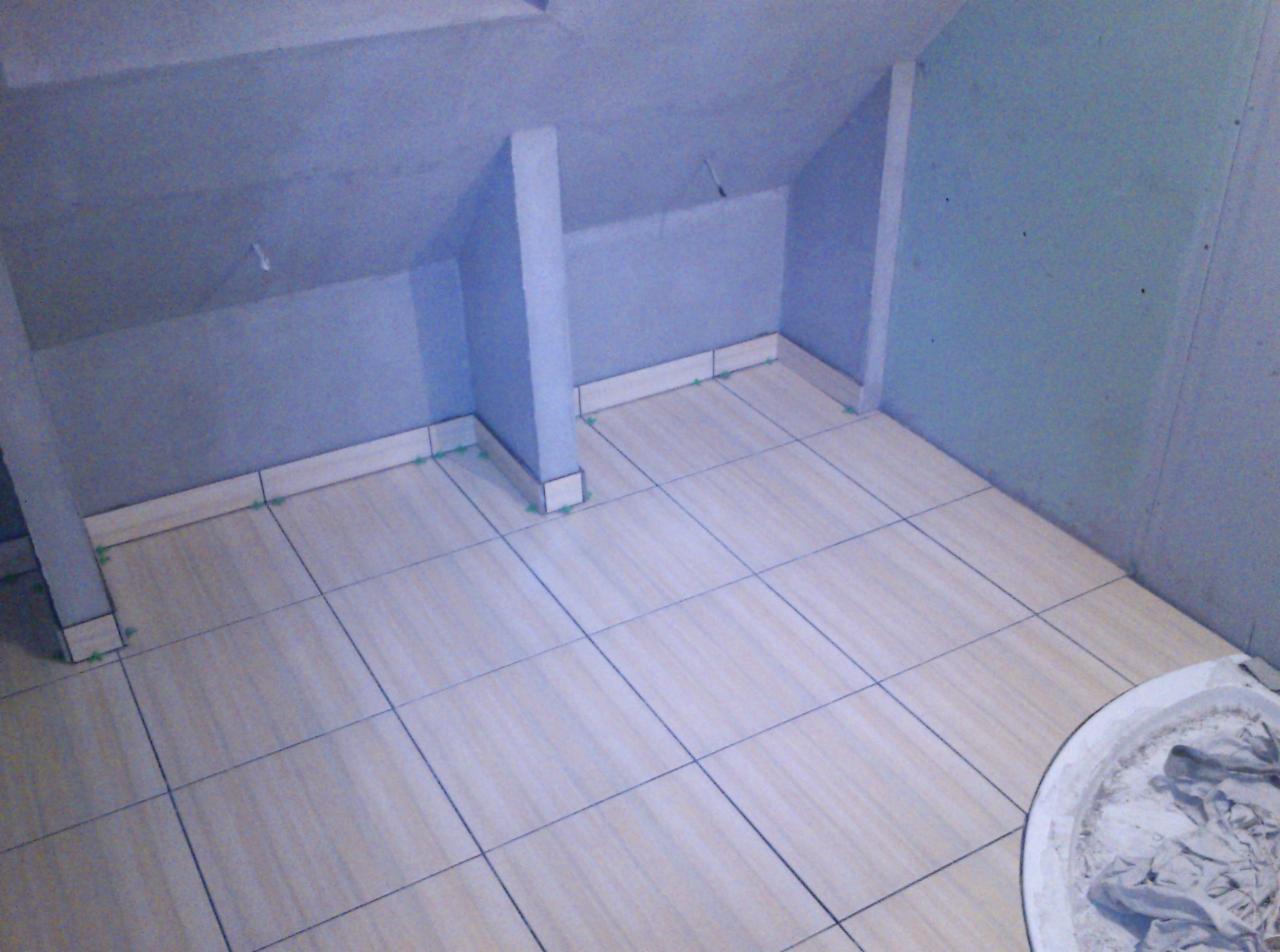 Wyłożona jasnymi płytkami podłoga na poddaszu. Widoczne szpachlowane ściany i słupki, przygotowane do dalszych prac wykończeniowych. Na podłodze widoczny pojemnik z zaprawą.