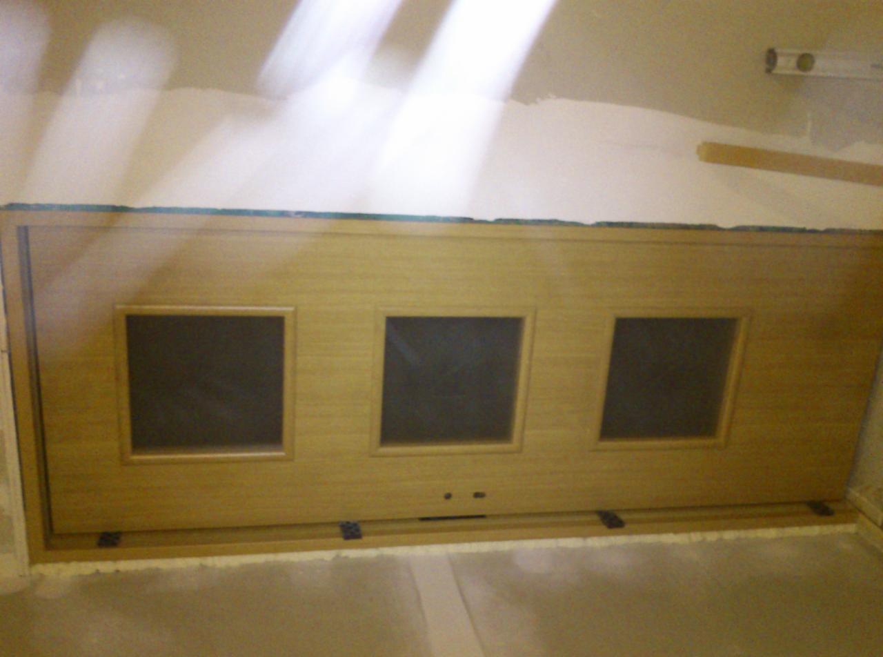 Drewniana zabudowa z trzema kwadratowymi otworami, osadzona w ścianie podczas prac wykończeniowych. Widoczne zawiasy i niedokończone wykończenie.