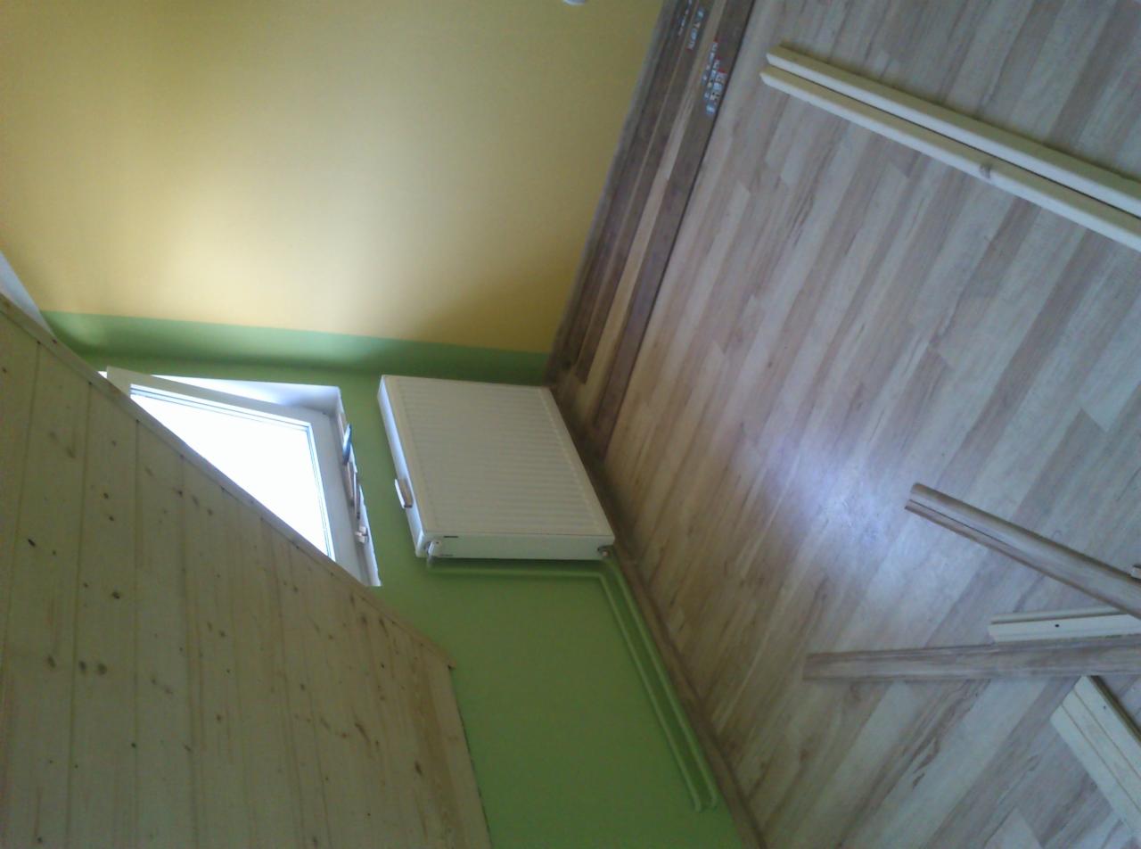 Pokój w trakcie remontu: drewniana boazeria, grzejnik na zielonej ścianie, okno, panele podłogowe oraz fragmenty listew przypodłogowych. Żółty sufit.