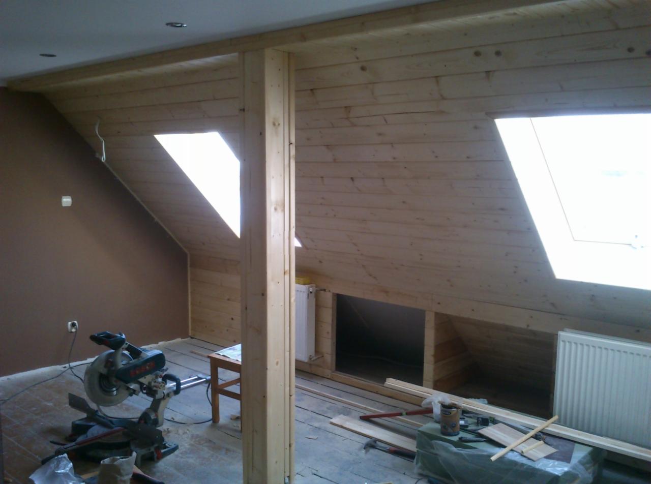 Drewniane wykończenie poddasza z oknami dachowymi, widoczna piła do drewna i narzędzia, etap prac wykończeniowych.