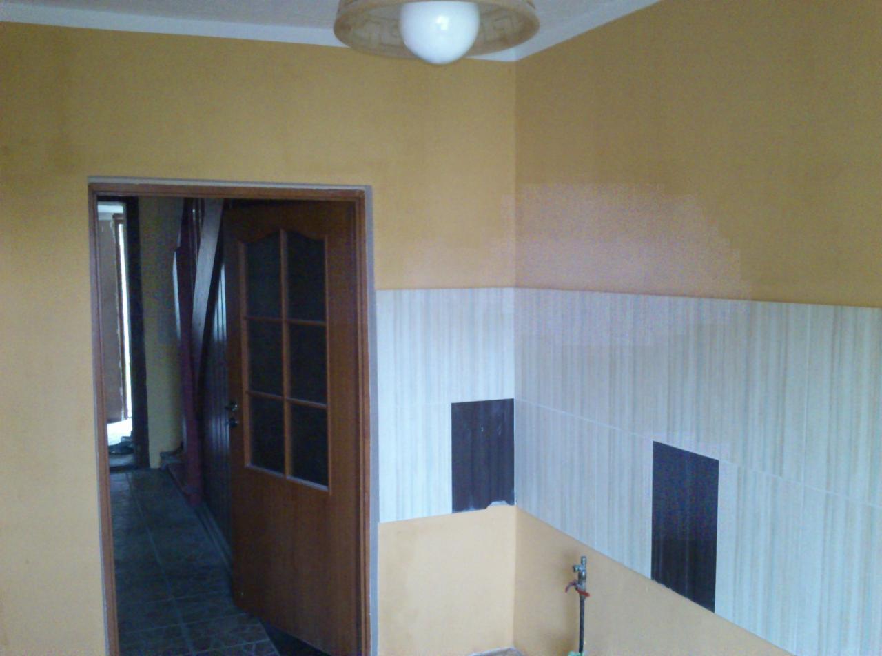 Żółta ściana z białymi płytkami i ciemnymi wstawkami w łazience. Częściowo widoczne drzwi z szybkami. W tle widoczny korytarz z podłogą w kratkę.