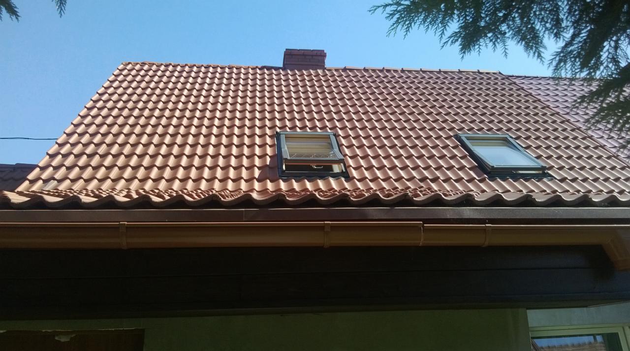 Czerwony dach z dachówki ceramicznej z dwoma oknami dachowymi, jedno otwarte, brązowe rynny, komin i fragment drzewa. Błękitne niebo w tle.