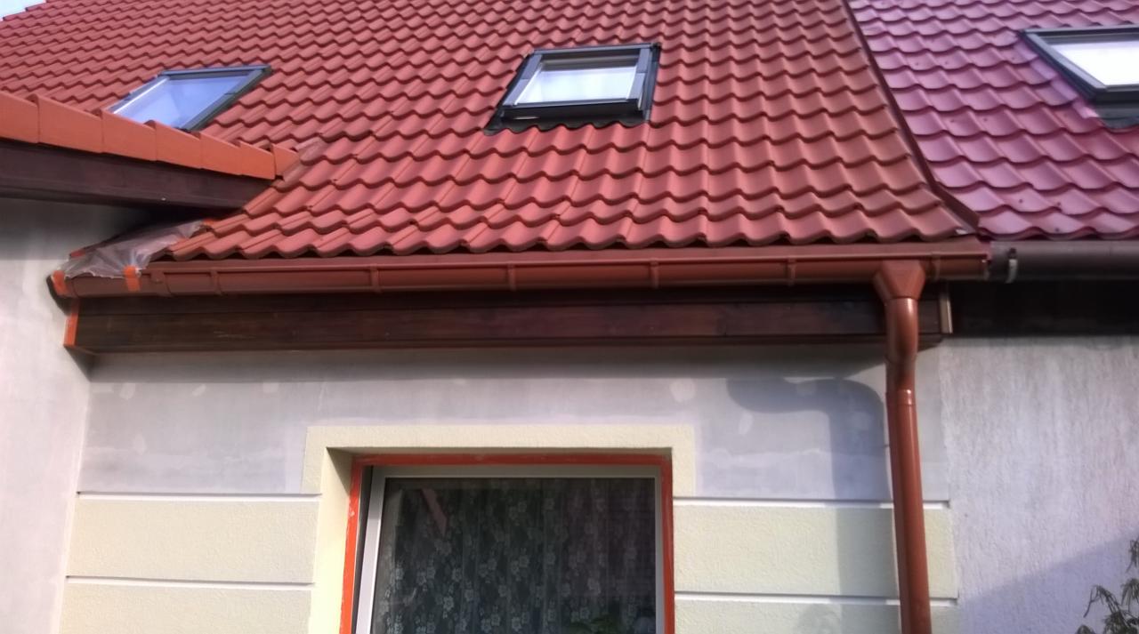 Dom z czerwoną dachówką, brązowymi rynnami i oknem. Elewacja częściowo otynkowana, z widocznymi detalami architektonicznymi. Dach wyposażony w okna dachowe.