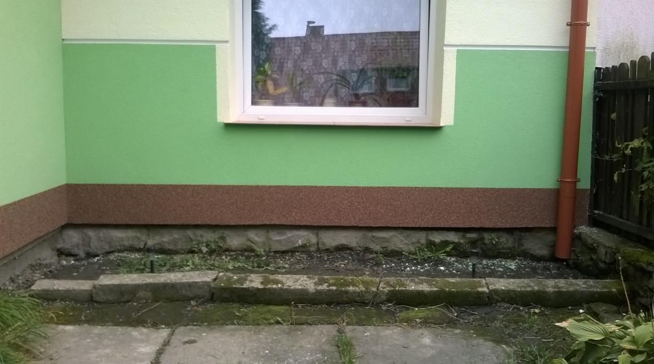 Fragment elewacji domu z oknem i rynną, pomalowany w odcieniach zieleni i brązu, z widocznym fundamentem i fragmentem ogrodzenia.