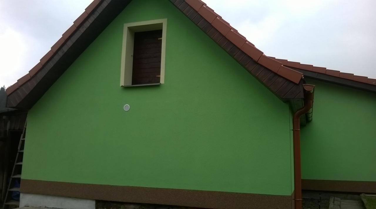 Ściana domu po malowaniu na zielono z brązowymi elementami dachu i rynny. Widoczny fragment drabiny przy ścianie.