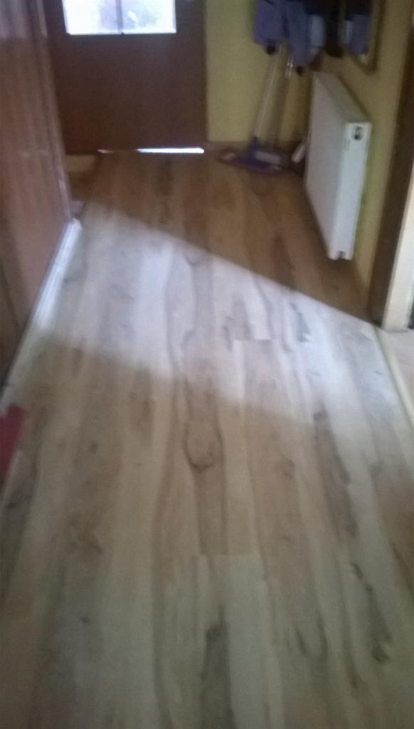 Jasne panele podłogowe w przedpokoju, widoczne drzwi wejściowe i fragment grzejnika. Ujęcie z góry, podkreślające wzór paneli.