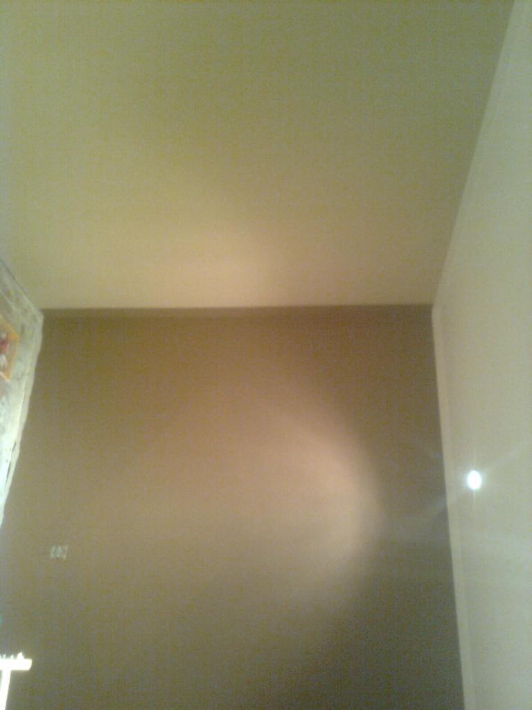 Gładka, brązowa ściana po tynkowaniu, widok z dołu, z fragmentem sufitu i narożnika. Widoczne niedoskonałości oświetlenia.