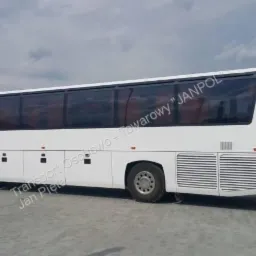 Biały autokar turystyczny z przyciemnianymi szybami, zaparkowany na szarym placu, widok z boku.