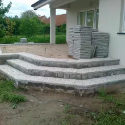 Wykończone schody wejściowe z szarej kostki, przylegające do białego budynku. Obok ułożone stosy kostki, teren wokół częściowo zagospodarowany.