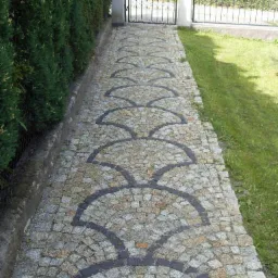 Ścieżka z jasnoszarej kostki granitowej ułożonej w dekoracyjny wzór wachlarzy, prowadząca do furtki z czarnego metalu, otoczona zielenią.