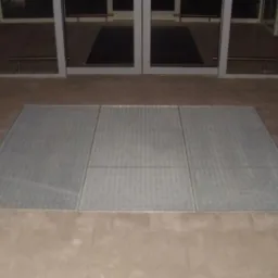 Metalowa krata wkomponowana w posadzkę przed wejściem z przeszklonymi drzwiami. Widoczny fragment ceglanej ściany po prawej stronie.