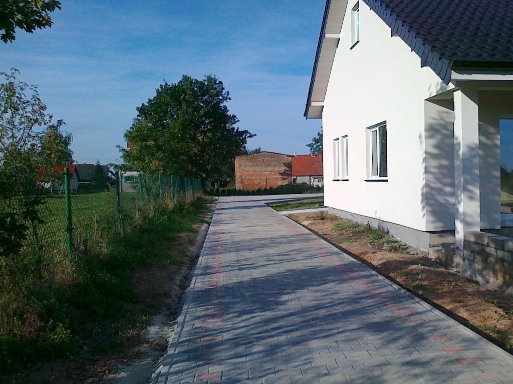 Ułożona, szara kostka brukowa z czerwonymi akcentami tworzy ścieżkę wzdłuż białego domu z brązowym dachem. Widoczne drzewa i ogrodzenie w tle.
