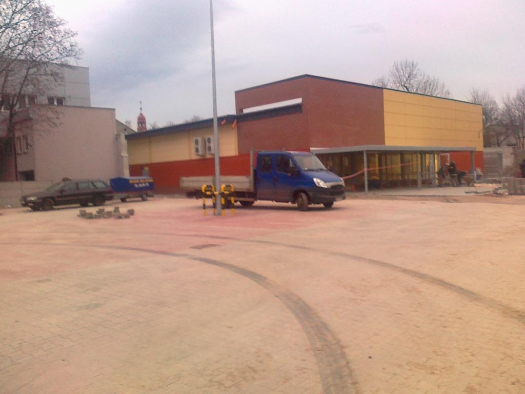 Nierówna nawierzchnia z kostki, ślady opon, niebieska ciężarówka na tle budynku z żółto-pomarańczową elewacją i fragmentem szarego nieba. Widoczne zaparkowane auto i przyczepa.