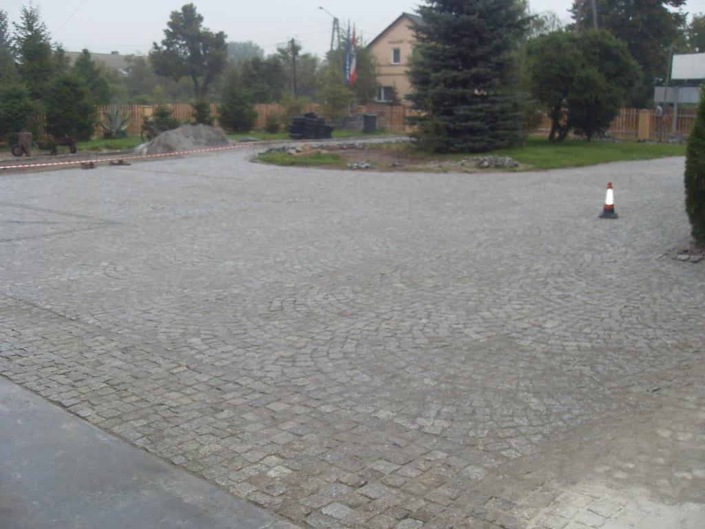 Szara kostka granitowa ułożona w łukowaty wzór na placu, z pomarańczowym pachołkiem i elementami krajobrazu w tle, w pochmurny dzień.