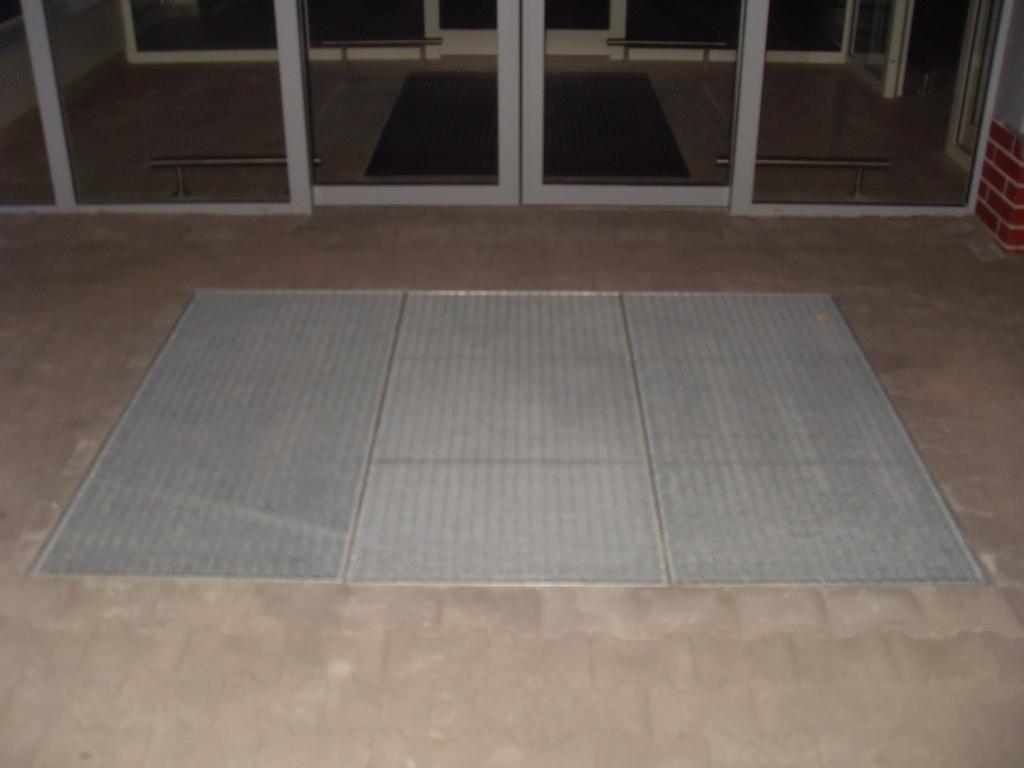 Metalowa krata wkomponowana w posadzkę przed wejściem z przeszklonymi drzwiami. Widoczny fragment ceglanej ściany po prawej stronie.