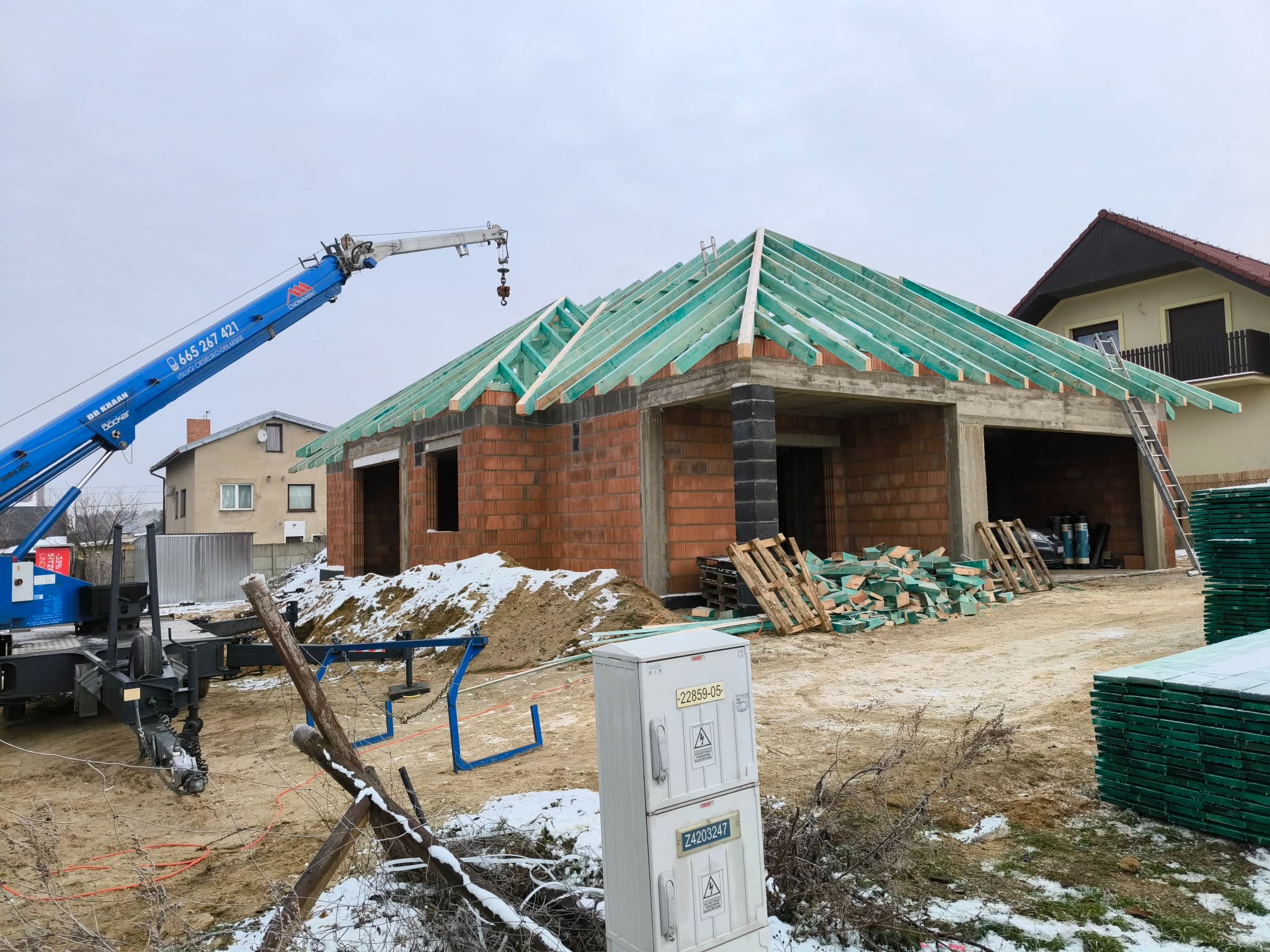 Budowa domu jednorodzinnego w trakcie: murowane ściany, więźba dachowa z zielonych łat, dźwig budowlany w tle. Częściowo ośnieżony teren budowy.