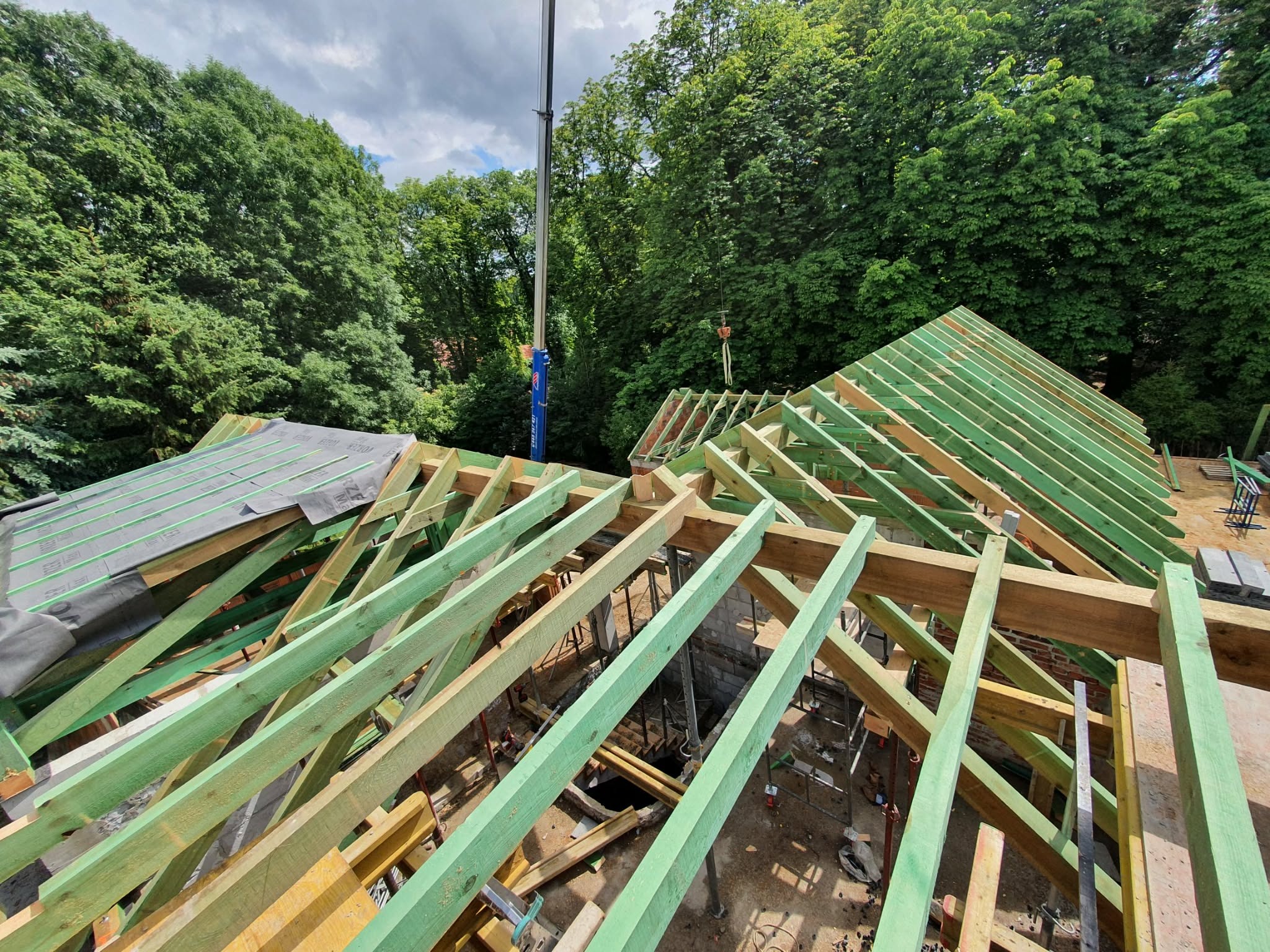 Wysoki kąt: konstrukcja dachu z zielonych i brązowych belek, widoczny dźwig w tle, otoczona zielenią drzew. Etap budowy więźby dachowej z bliska.