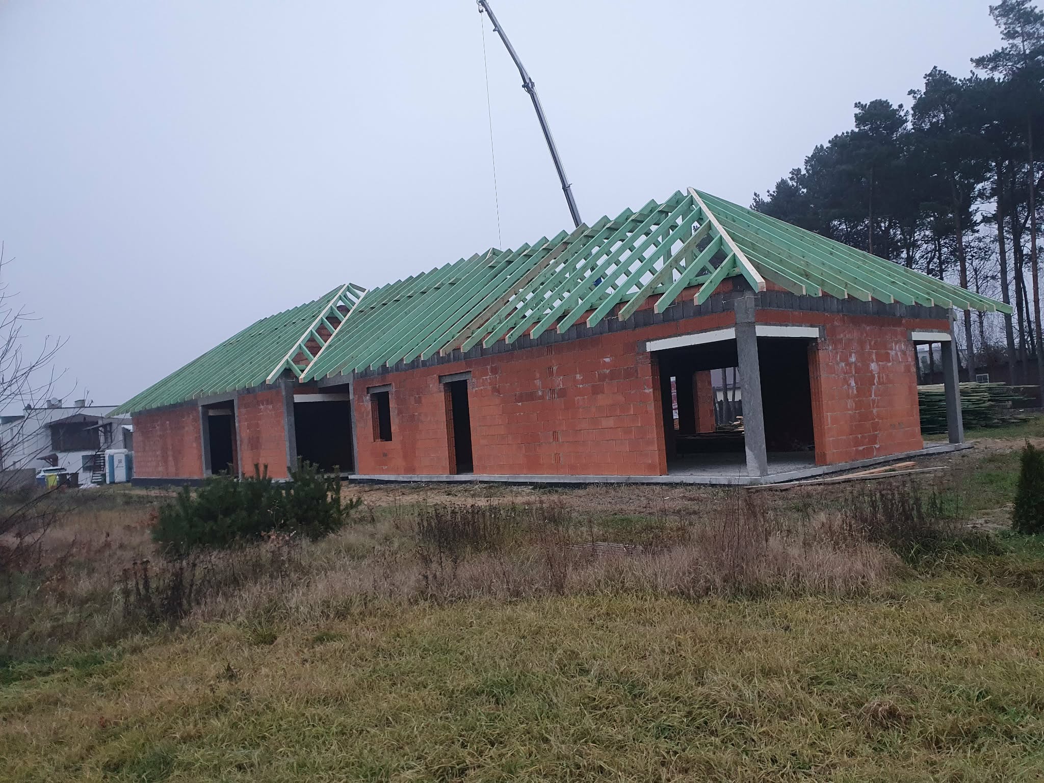 Surowy stan budynku z cegły z zieloną więźbą dachową w trakcie montażu, widoczny żuraw budowlany w tle, pochmurny dzień.