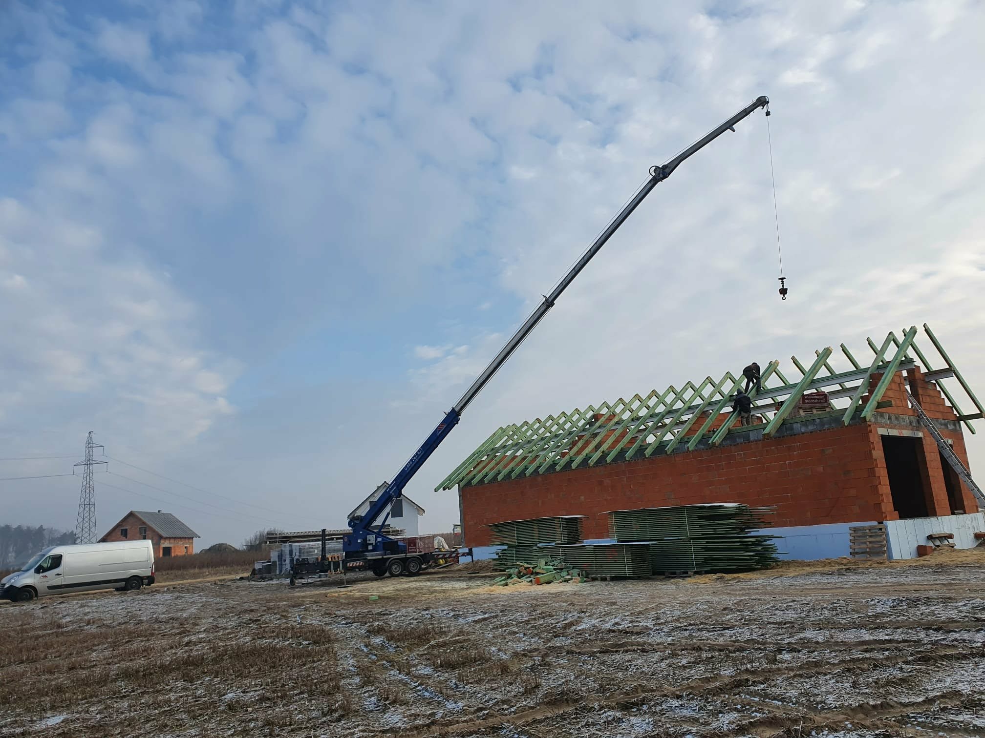 Budowa domu jednorodzinnego: mur z czerwonej cegły, zielona więźba dachowa w trakcie montażu z użyciem dźwigu. W tle krajobraz z zabudowaniami i pochmurne niebo.