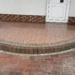 Półokrągłe wejście do domu wyłożone czerwoną kostką brukową, oddzielone od chodnika obrzeżem z ciemnobrązowej kostki i jasnego grysu, z widoczną białą kolumną i drzwiami.