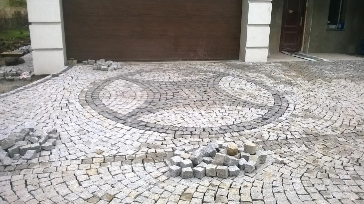 Wykończony podjazd z kostki granitowej ułożonej w dekoracyjny wzór koncentrycznych okręgów, z elementami do ułożenia w pobliżu, brama garażowa w tle.