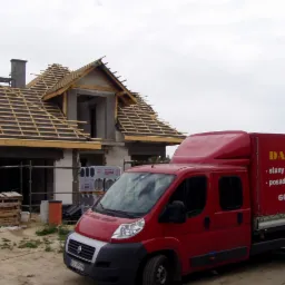 Budowa domu w stanie surowym otwartym z widoczną konstrukcją dachu i czerwonym samochodem dostawczym firmy budowlanej na pierwszym planie.