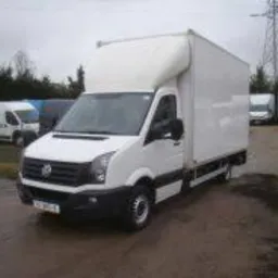 Biały samochód dostawczy z wysoką zabudową na placu, widoczne inne pojazdy w tle, pochmurne niebo.