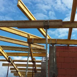 Szczegółowy widok szalunku stropu z belek drewnianych i podpór stalowych na tle błękitnego nieba z chmurami, widoczne fragmenty muru z czerwonej cegły.