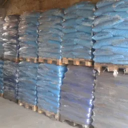 Magazyn z paletami ułożonymi w stosy, na których znajdują się niebieskie worki z napisem. W tle widoczny wózek widłowy.