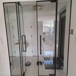 FHU Inny Wymiar Szkła Justyna Ługowska - Szklane drzwi loft z czarnymi ramami, widok na wnętrze domu. Na podłodze leżą buty, a w tle widać wejście i oświetlenie sufitowe.