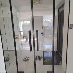 FHU Inny Wymiar Szkła Justyna Ługowska - Szklane drzwi loft z czarnymi ramami i podłużnymi uchwytami, widoczne buty w przedpokoju, marmurowa podłoga i kryształowy żyrandol w tle.