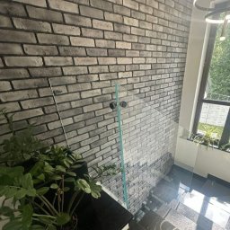 FHU Inny Wymiar Szkła Justyna Ługowska - Szklana balustrada zabezpiecza schody z czarnego kamienia, na tle ściany z cegły w odcieniach szarości. W tle widoczne okno z roślinami.