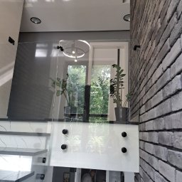 FHU Inny Wymiar Szkła Justyna Ługowska - Szklana balustrada schodowa z czarnymi mocowaniami, widok z dołu. Fragment ściany z cegły, rośliny doniczkowe i schody z marmurowym wzorem.