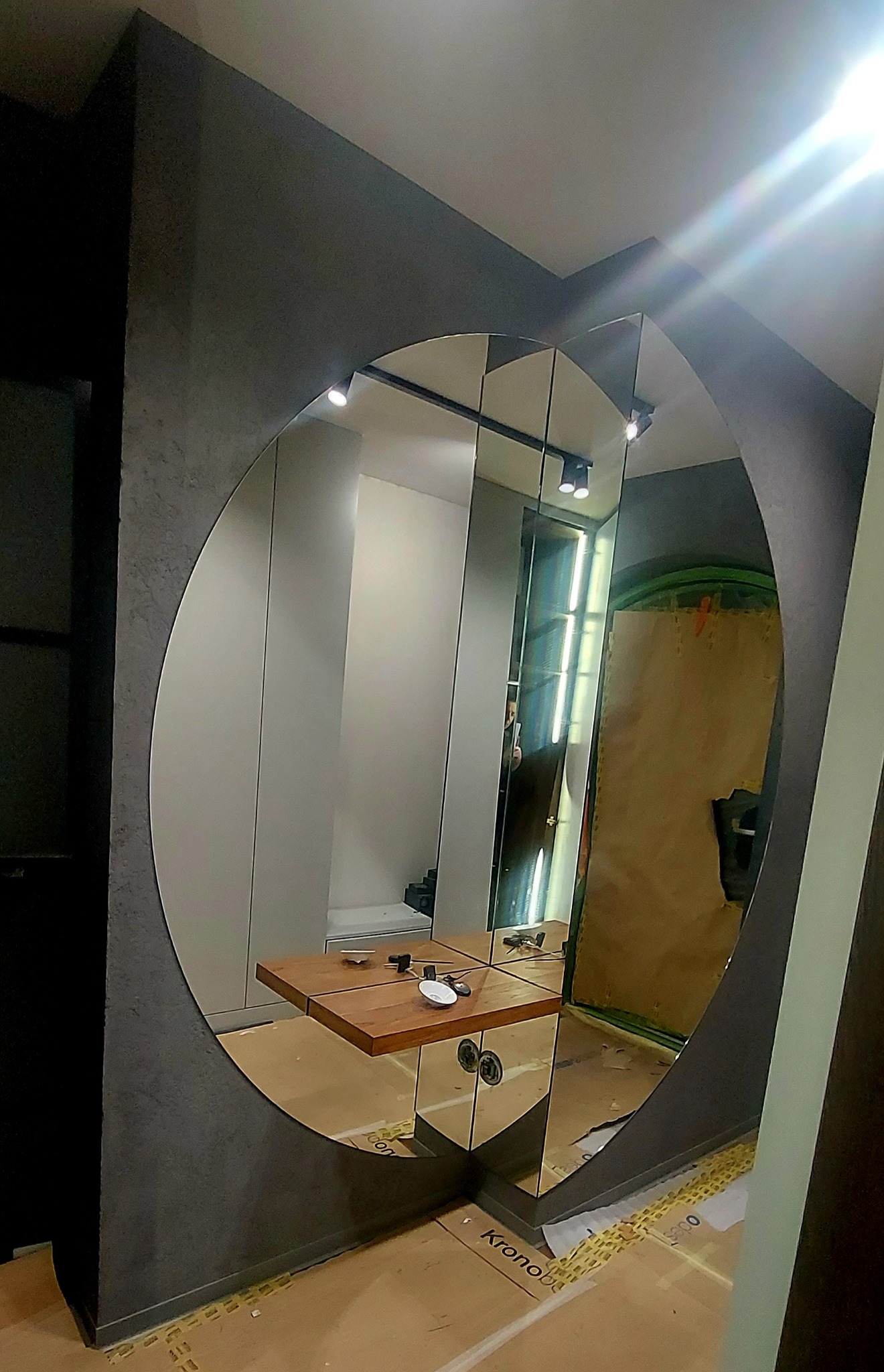 Okrągłe lustro w szarej ramie, odbijające fragment wnętrza z drewnianym blatem i kartonami na podłodze.