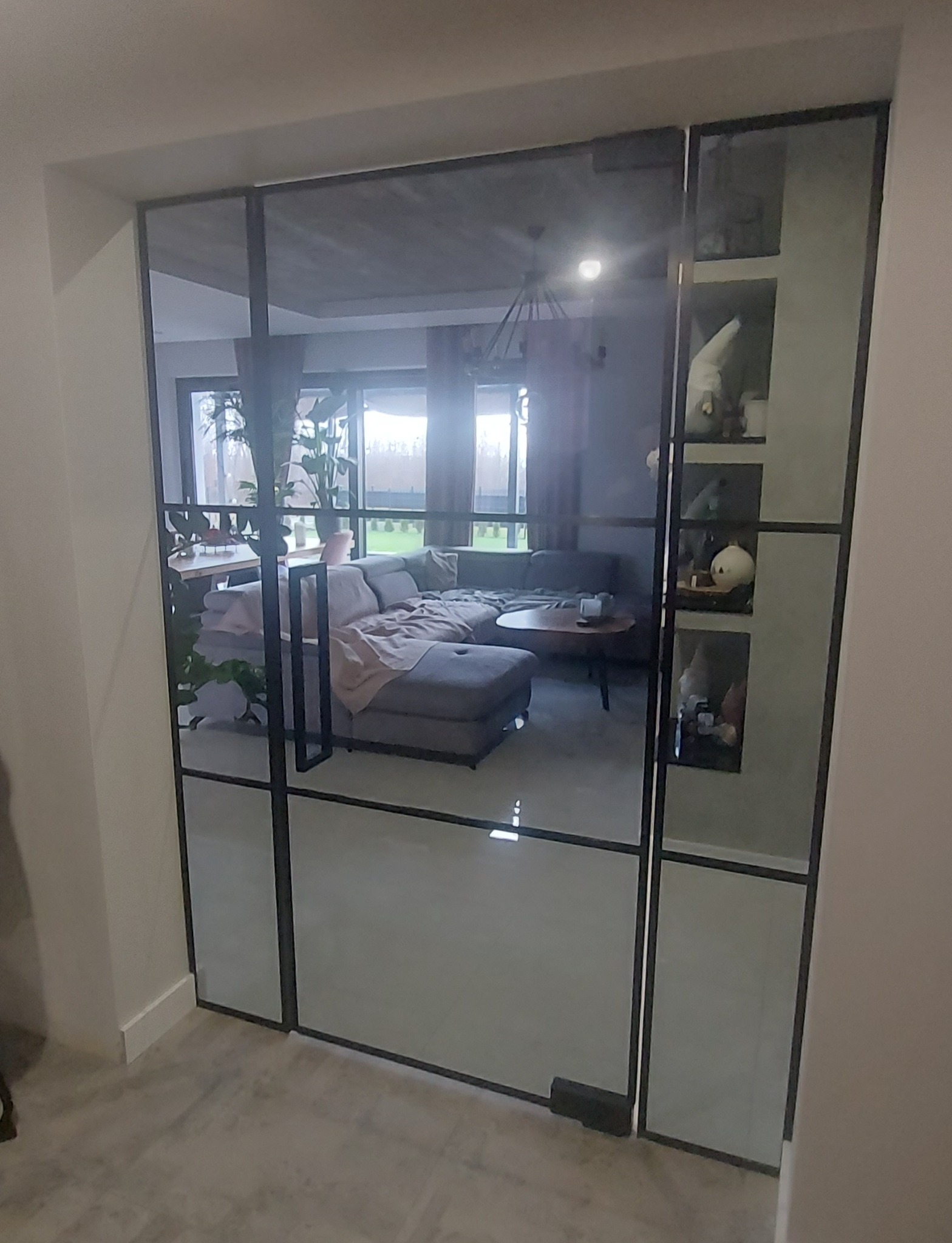 Szklana ścianka działowa z czarnymi profilami oddzielająca pokój dzienny z szarą sofą i dekoracyjnymi figurkami na półkach.