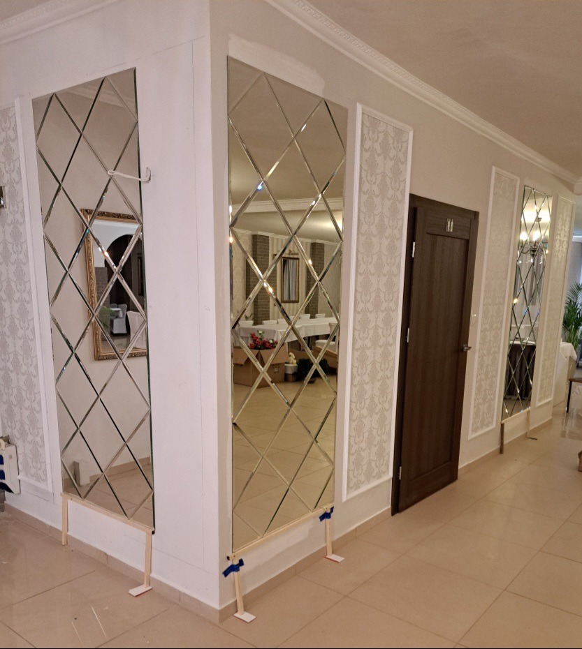 Montaż mozaiki lustrzanej z romboidalnych elementów na ścianie, zabezpieczonej drewnianymi podpórkami podczas instalacji, widoczne odbicie sali bankietowej.