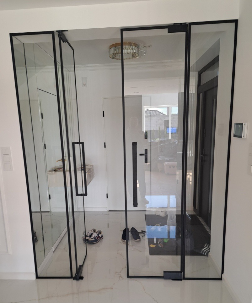 Szklane drzwi loft z czarnymi ramami, widok na wnętrze domu. Na podłodze leżą buty, a w tle widać wejście i oświetlenie sufitowe.