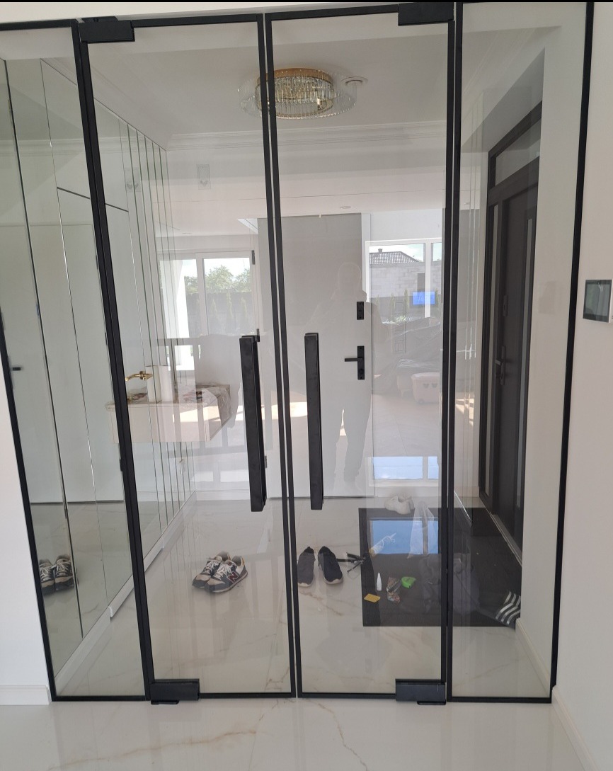 Szklane drzwi loft z czarnymi ramami i podłużnymi uchwytami, widoczne buty w przedpokoju, marmurowa podłoga i kryształowy żyrandol w tle.