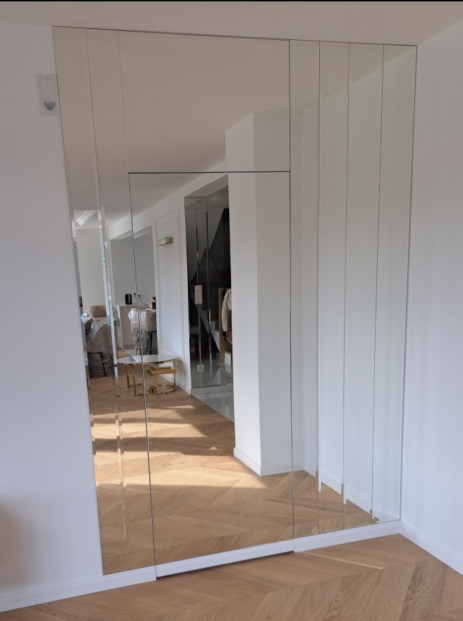 Ściana pokryta mozaiką z pionowych, lustrzanych paneli, odbijających wnętrze pokoju z parkietem ułożonym we wzór jodełki. Widoczny fragment białej ściany i listwy przypodłogowej.