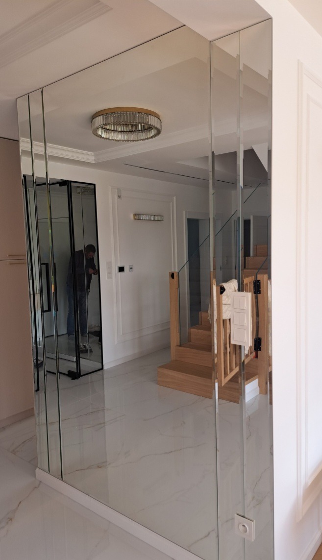 Eleganckie lustro ścienne z fazowanych paneli w nowoczesnym wnętrzu. Widoczne odbicie schodów i drzwi przesuwnych, podkreślające przestronność i unikalny design.