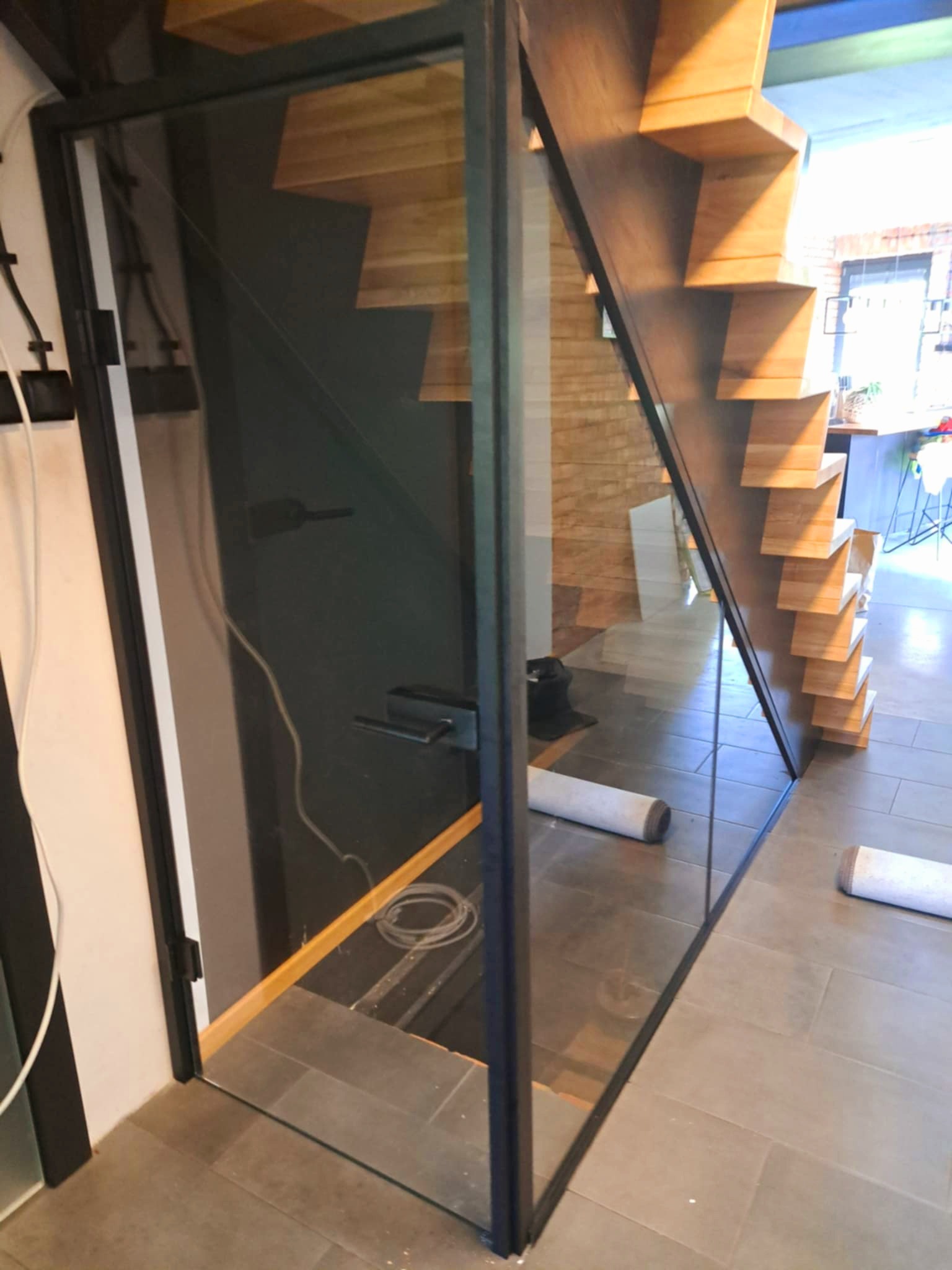 Nowoczesna, szklana balustrada schodowa z czarną ramą, zabezpieczająca drewniane schody o ażurowej konstrukcji. Widoczne elementy montażowe i fragment zwiniętego dywanu.