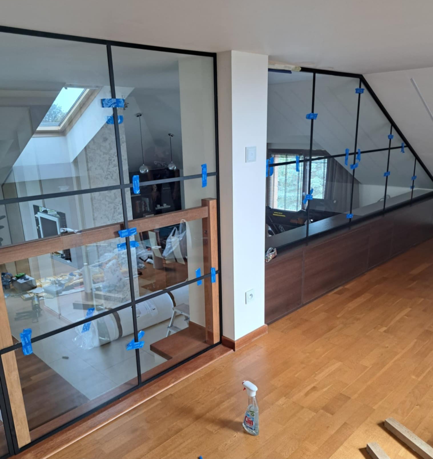 Szklana balustrada z czarnymi profilami, zabezpieczona niebieskimi klamrami montażowymi, oddzielająca przestrzeń na poddaszu od schodów. Widok na drewnianą podłogę i butelkę ze środkiem czyszczącym.