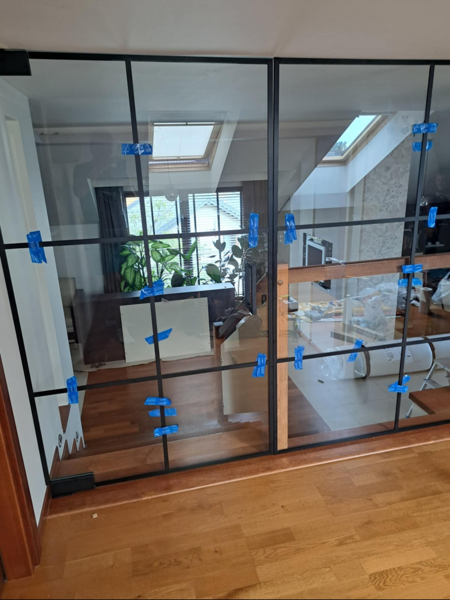 Szklana ścianka działowa z czarnymi profilami, zabezpieczona niebieską taśmą, oddzielająca przestrzeń wewnątrz domu. Widoczne schody i okno dachowe.