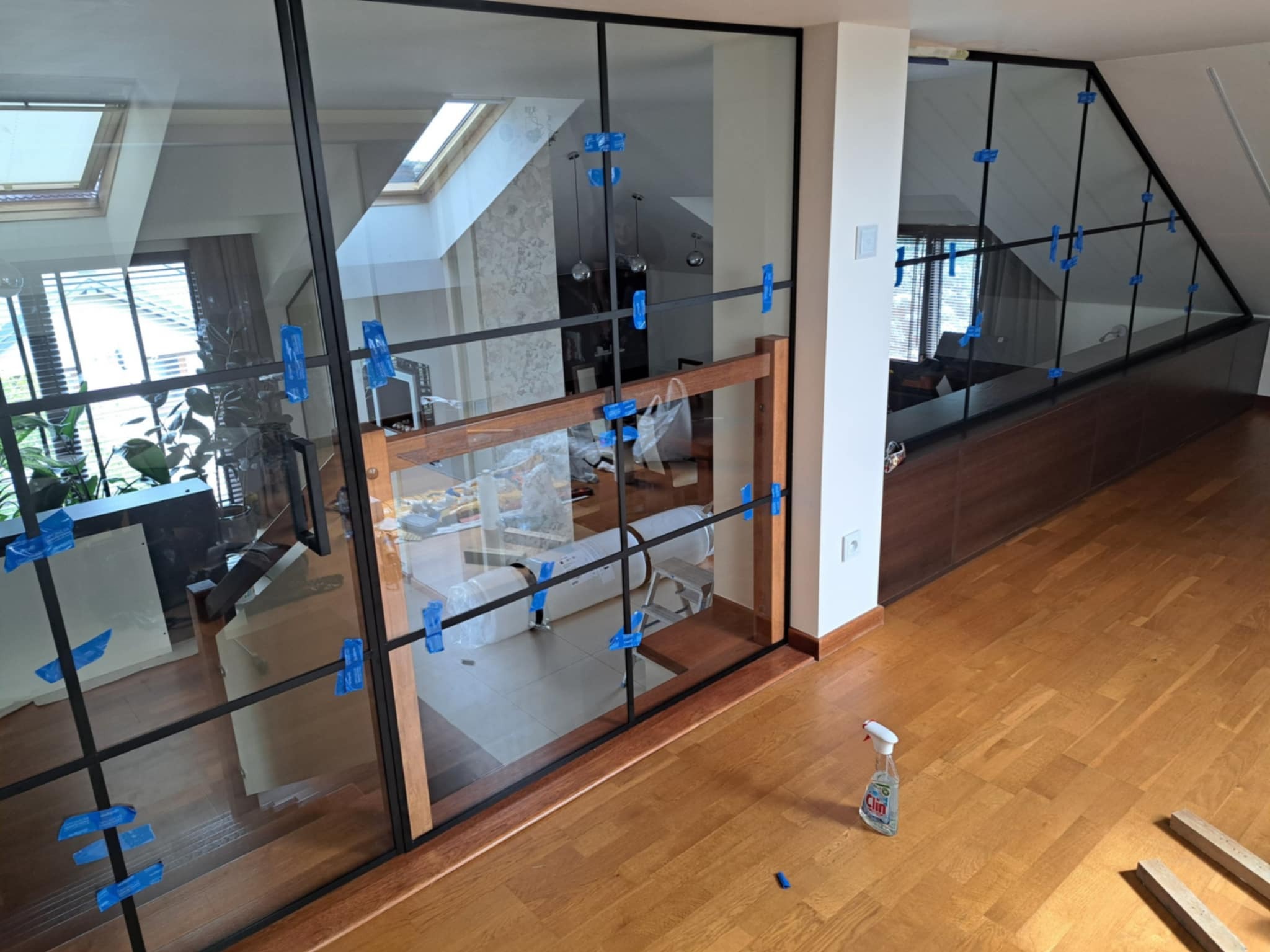 Szklana konstrukcja z czarnymi profilami i drewnianymi elementami, zabezpieczona niebieskimi klipsami montażowymi, na tle jasnego wnętrza z oknami dachowymi.