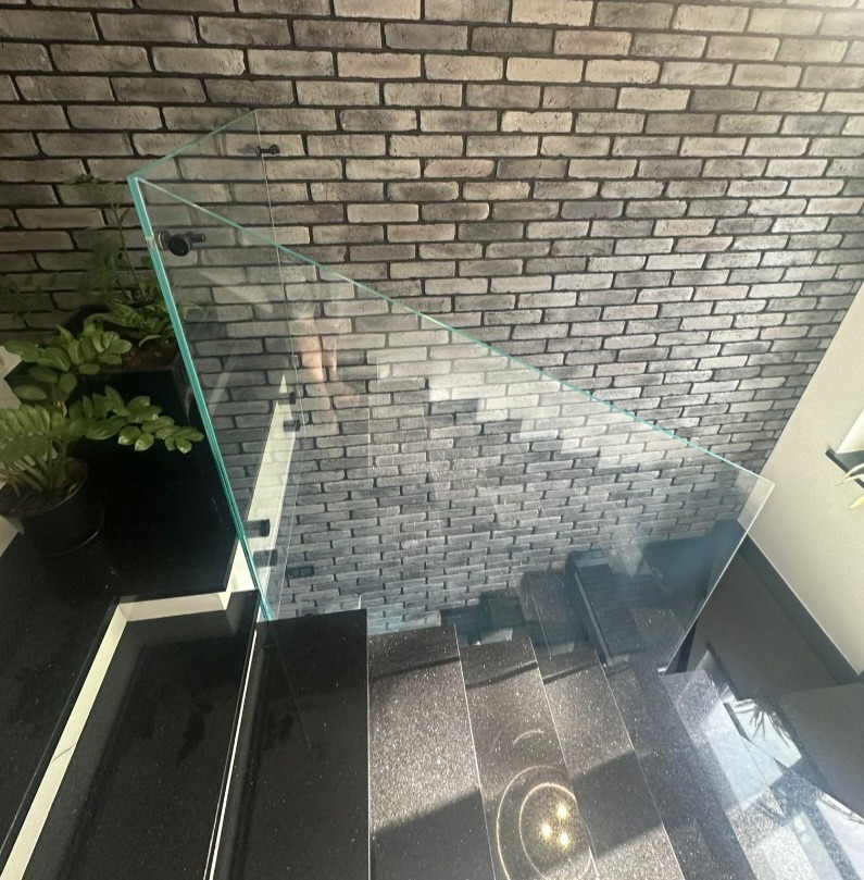 Szklana balustrada schodowa z mocowaniem punktowym na tle ściany z szarej cegły i czarnych schodów. Minimalistyczny design, nowoczesne wykończenie.