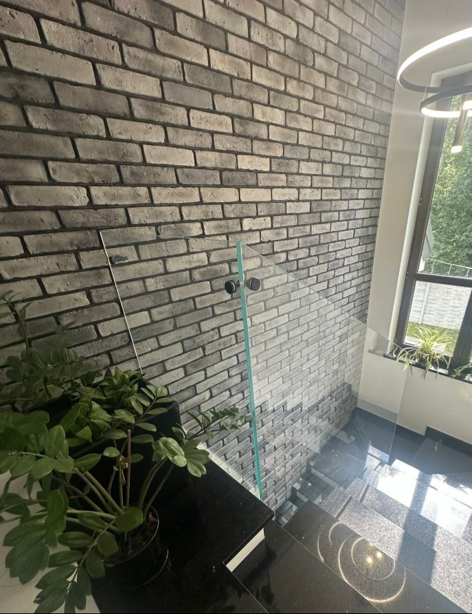 Szklana balustrada zabezpiecza schody z czarnego kamienia, na tle ściany z cegły w odcieniach szarości. W tle widoczne okno z roślinami.
